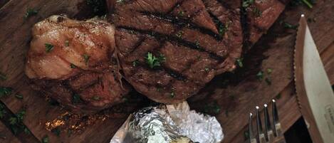 A wooden cutting board with a steak, vegetables, and foil-wrapped potatoes.