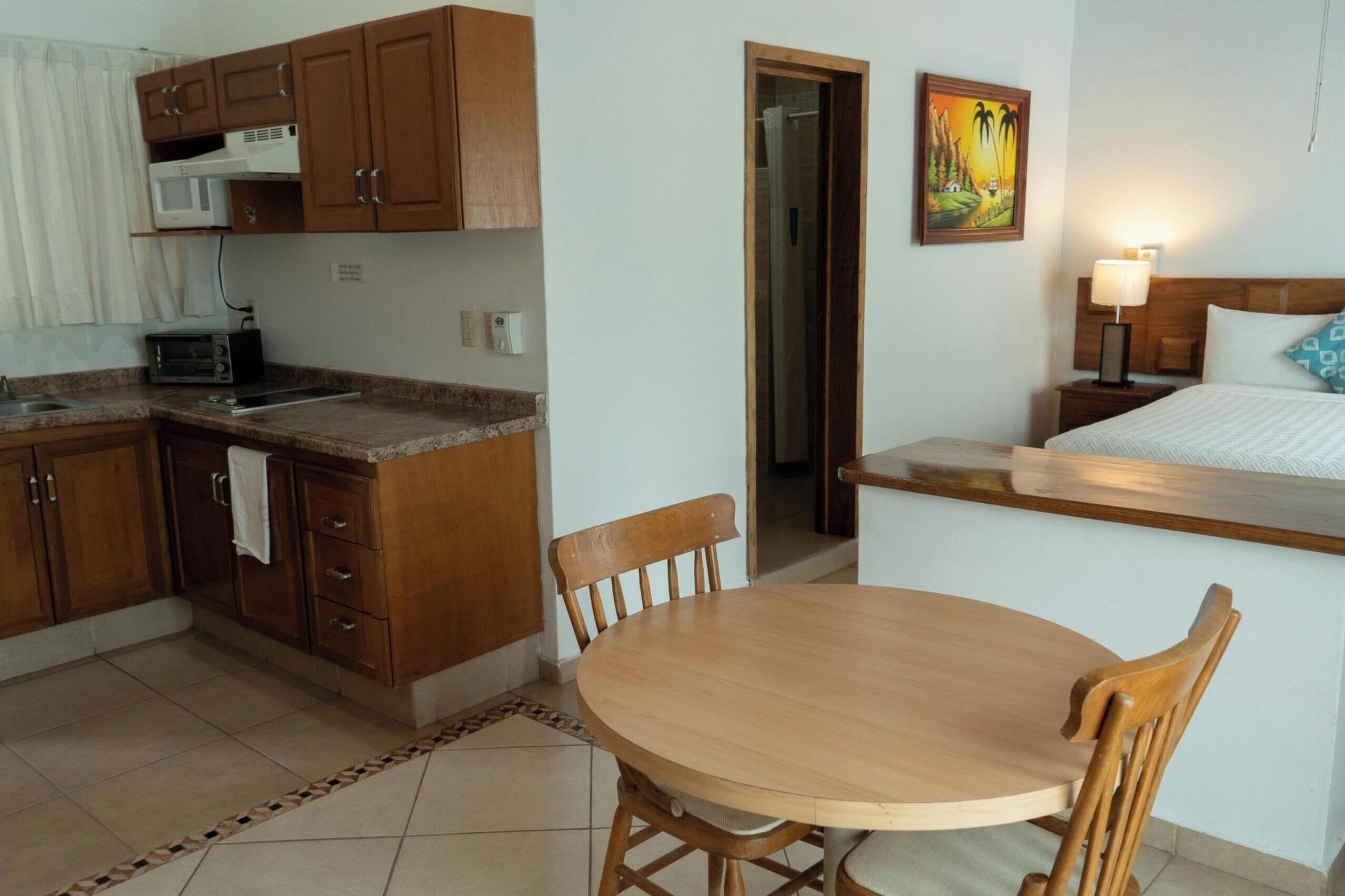 Habitación de hotel compacta con kitchenette, comedor con mesa y sillas, cama con cabecera y un cuadro en la pared.