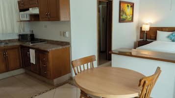 Habitación de hotel compacta con kitchenette, comedor con mesa y sillas, cama con cabecera y un cuadro en la pared.