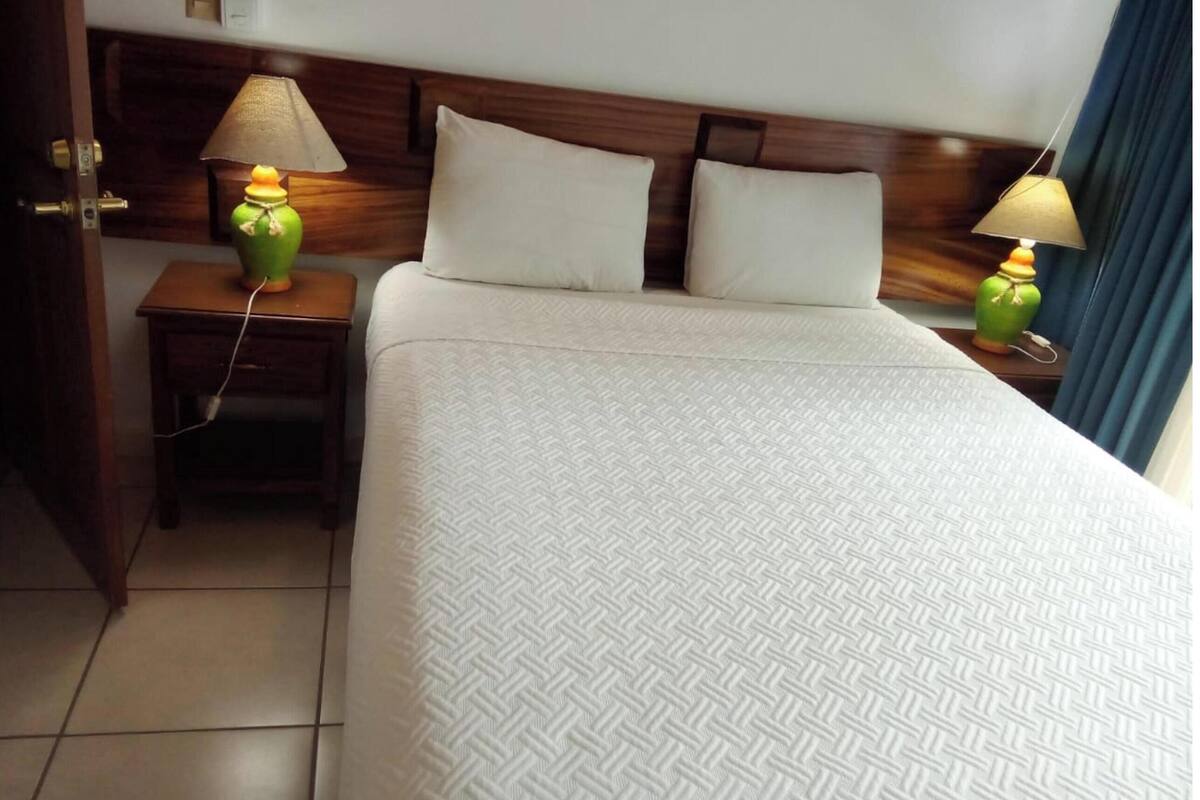 A hotel room with a large bed, two bedside lamps, a wooden headboard, and a tiled floor.