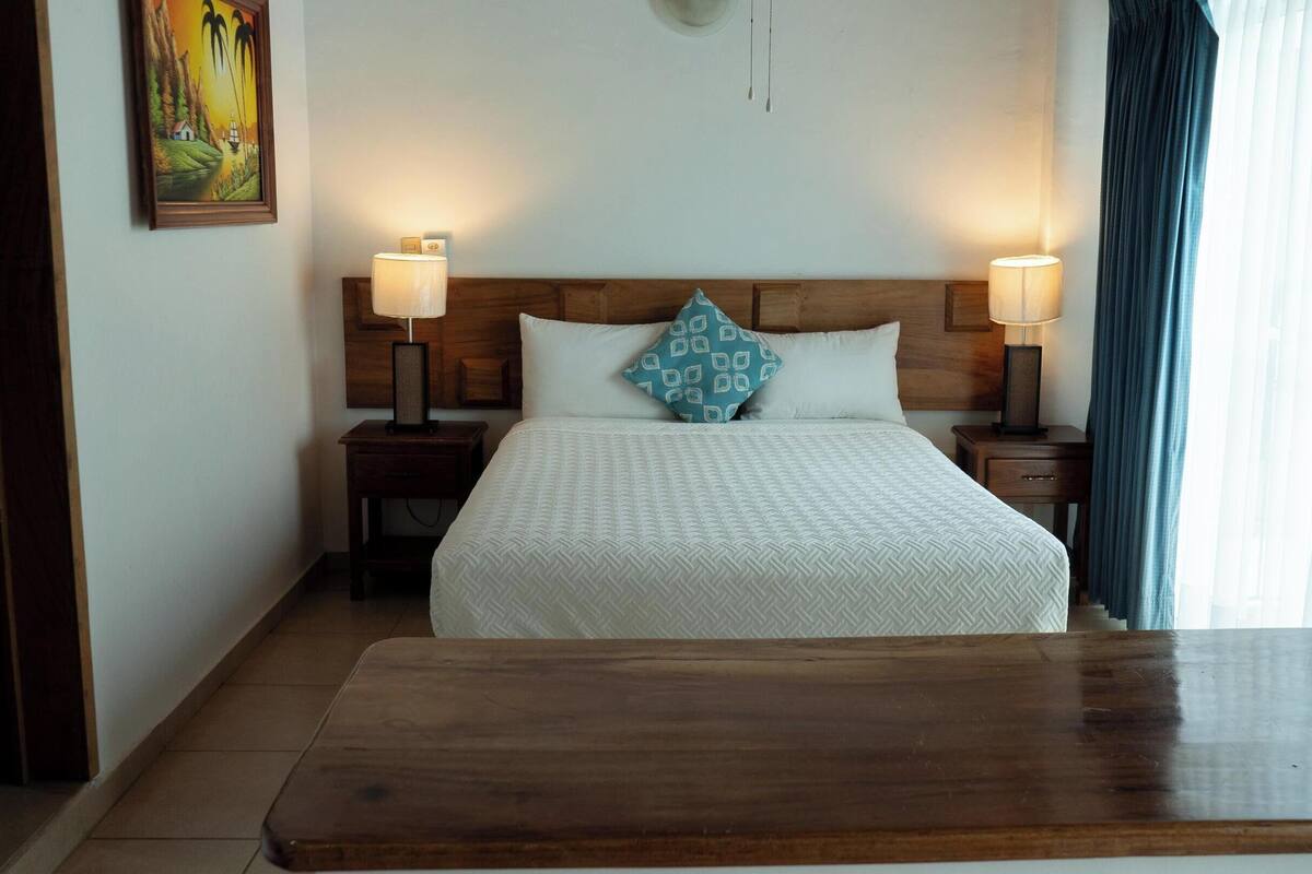 A hotel room with a wooden headboard, two bedside tables with lamps, a bed with a patterned bedspread, and a painting on the wall.