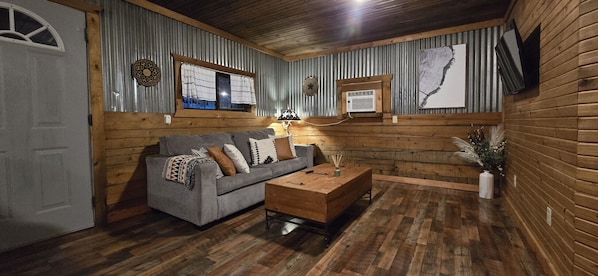 Une pièce chaleureuse avec un plancher en bois, un canapé gris agrémenté de coussins à motifs, une table basse en bois et un climatiseur fixé au mur.