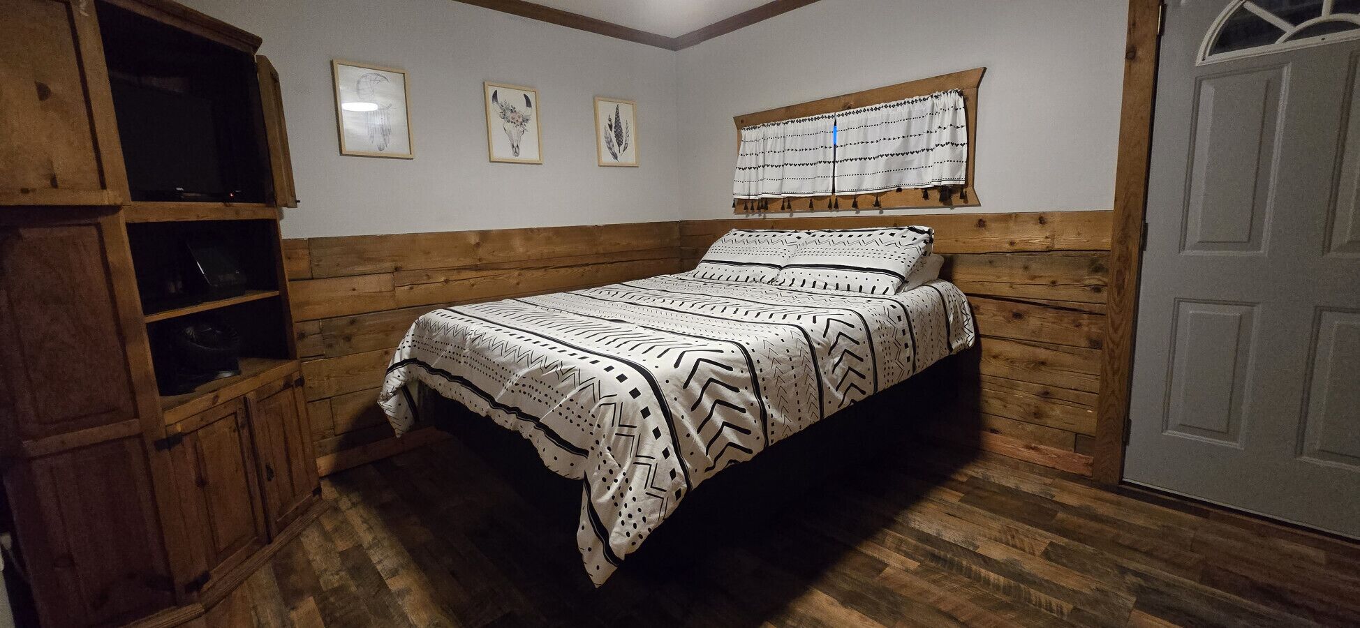 A bedroom with a bed, wooden walls, a window with curtains, and a door.