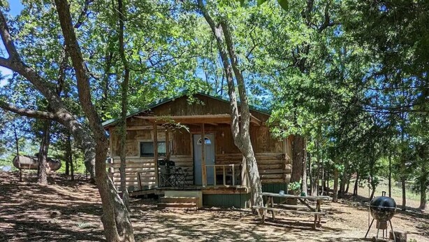A rustic cabin surrounded by trees, with a porch and a grill area.