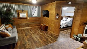 A cozy log cabin interior with a grey sofa, a small wooden table, a television mounted on the wall, and a bed with a white and patterned bedspread.