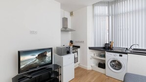 Cocina funcional con lavadora, microondas y estufa. Hay una ventana con persianas y un televisor de pantalla plana montado en la pared.