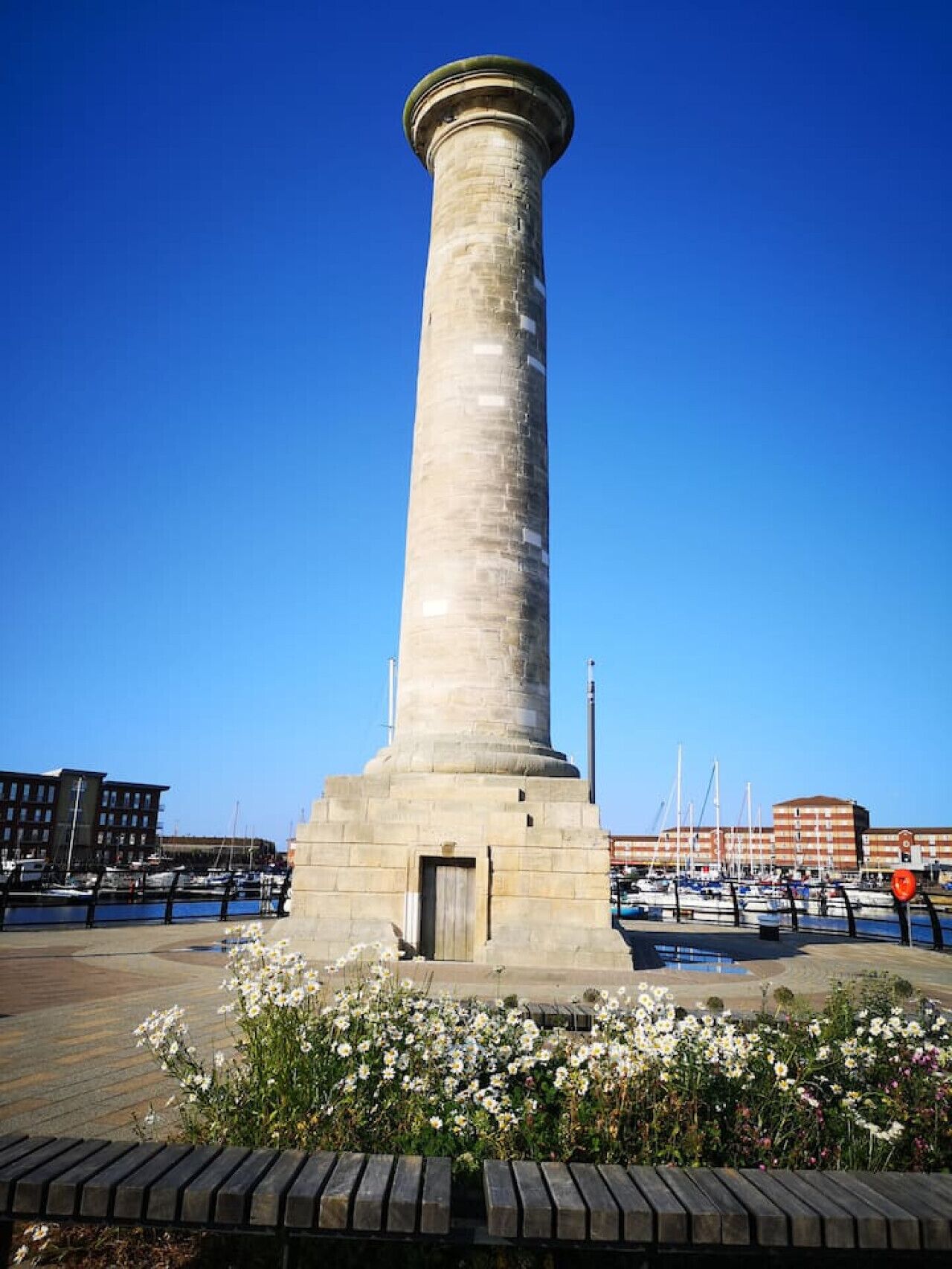 A tall, cylindrical stone monument with a door at the base, surrounded by a waterfront promenade with flowers and benches.