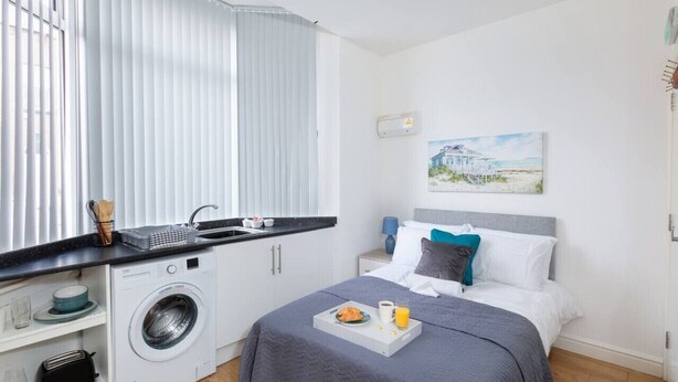 Una habitación de hotel moderna con una cama, una kitchenette pequeña, una lavadora y un cuadro en la pared.