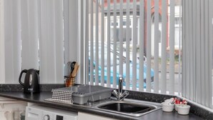 Una cocina con un hervidor negro, un lavamanos y una ventana con persianas verticales.