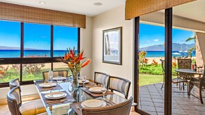 A dining area with a table set for four, large windows offering a view of the ocean, and a framed picture on the wall. - Maui Resort Rentals: Kaanapali Alii 315 –  2BR Oceanfront Ground-Floor Residence, Stunning Remodel, Mere Steps to Pools, Beach, & BBQs! (Lahaina)
