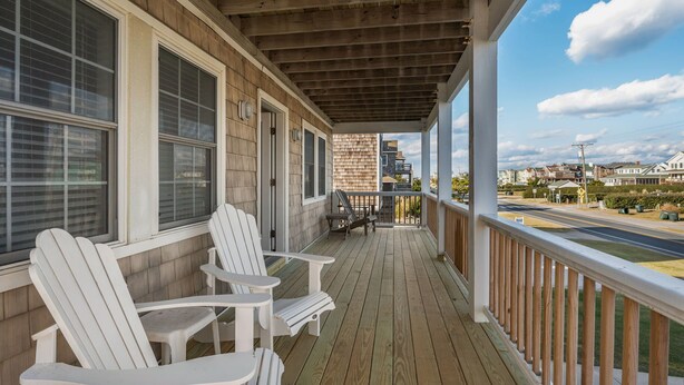 A covered porch with white Adirondack chairs, a table, and a view of a cityscape. - Pelican's Post: Semi-Oceanfront, Private Pool, Hot Tub and Game Room (Nags Head)