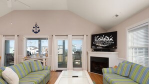 A living room with two striped sofas, a glass-top coffee table, a fireplace, and a TV displaying 'Coastal Carolina Vacation and Sales'. - Pelican's Post: Semi-Oceanfront, Private Pool, Hot Tub and Game Room (Nags Head)