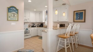 A kitchen with white cabinets, a central island with bar stools, a microwave, and a painting of a lighthouse. - Pelican's Post: Semi-Oceanfront, Private Pool, Hot Tub and Game Room (Nags Head)