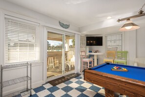 Uma mesa de bilhar com superfície em feltro azul, uma televisão presa à parede e uma porta que dá para área externa com vista dos arredores.