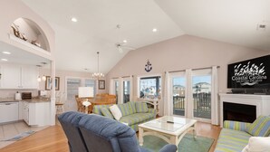 A spacious living area with a large sofa, a coffee table, and a television. There is a kitchen area with white cabinets and a dining table with chairs. - Pelican's Post: Semi-Oceanfront, Private Pool, Hot Tub and Game Room (Nags Head)