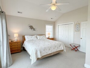 Ein Schlafzimmer mit Bett, Nachttisch, Lampe, Deckenventilator, Fenster mit Jalousien, Tür und Wanddekoration.