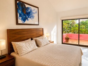 Quarto com uma cama grande, mesa de cabeceira com luminária, cabeceira de madeira e janela com vista para o verde.