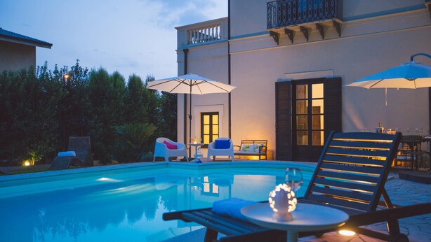 A swimming pool with lounge chairs and umbrellas, a dining table with chairs, and a building with a balcony. - Villa Afrodite 8 Emma Villas (Milazzo)
