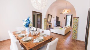 A dining area with a wooden table set for four, white chairs, and a vase of flowers. There is a white sofa and a television in the background. - Villa Afrodite 8 Emma Villas (Milazzo)