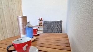Una mesa de madera con una taza y platillo rojos, una taza azul, un recipiente metálico y una silla de mimbre.