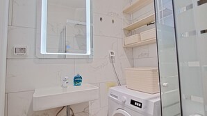Un baño moderno con pared de azulejos de mármol, lavamanos empotrado, lavadora y cabina de ducha con mampara de vidrio.