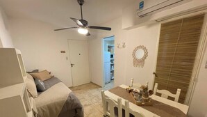 Une pièce de taille modeste comprenant un lit, une table à manger avec des chaises, un ventilateur de plafond et une porte donnant sur une autre pièce.