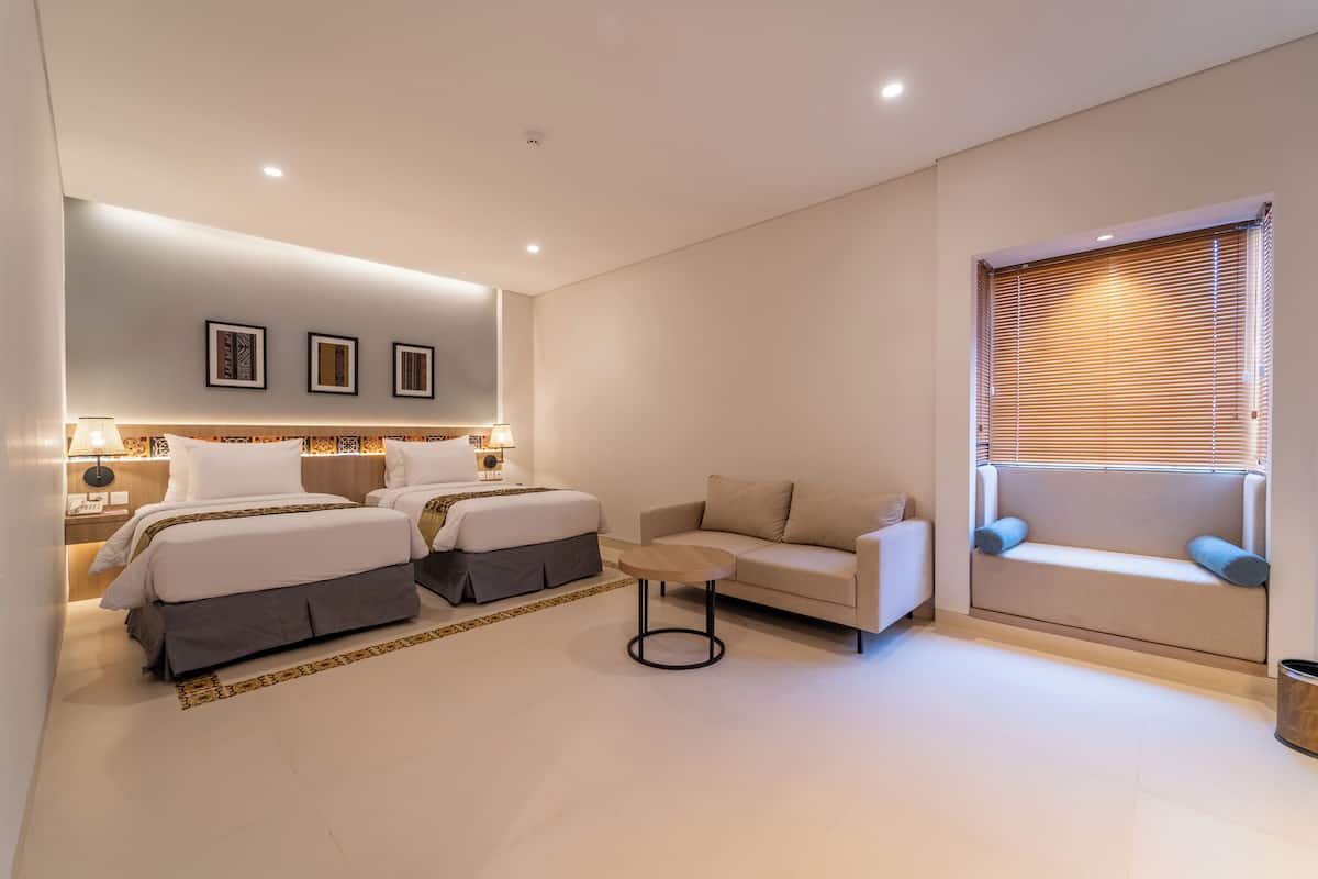 Une chambre d’hôtel moderne avec deux lits, un canapé, une petite table et une fenêtre avec des stores.