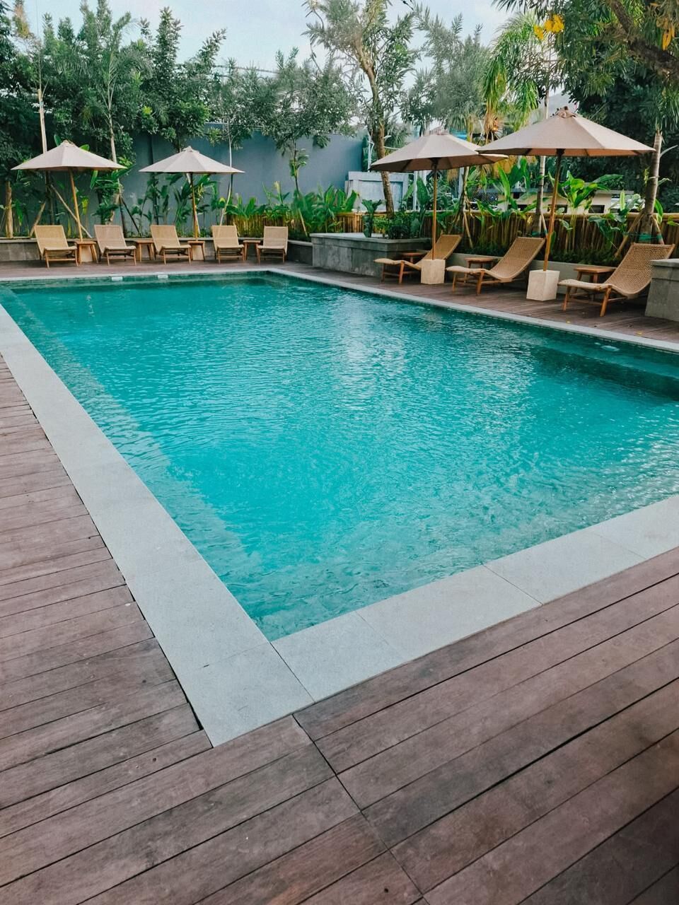 Piscina rectangular con suelo de madera y terrazas, rodeada de sillones y sombrillas.