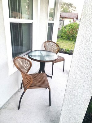 En uteplats med ett runt bord och flätade stolar, en vit vägg och en utsikt över ett hus i bakgrunden.