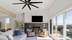 A modern living room with a fireplace, a ceiling fan, and a large window overlooking a balcony.