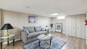 Ein Wohnzimmer mit grauem Sofa, einem Glastisch, einer Tischlampe mit Spitzenfächer, einem gerahmten Bild an der Wand und einem blauen Teppich mit Muster.