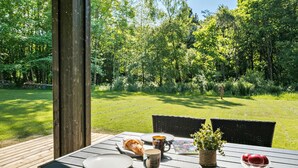Una terrazza esterna in legno con un tavolo apparecchiato per un pasto, circondata da una lussureggiante foresta verde.