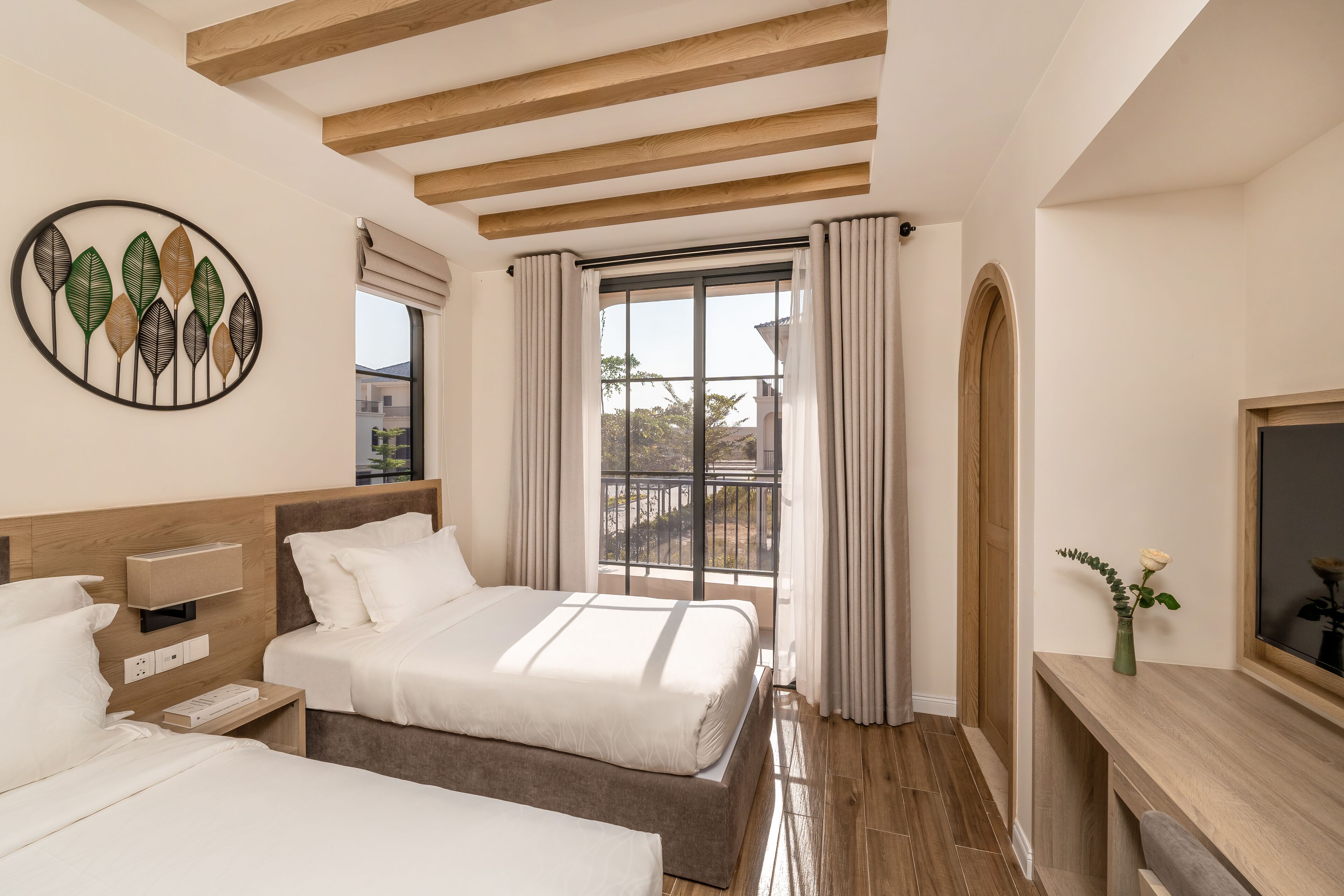 A hotel room with two beds, a wooden ceiling, a large window with curtains, a wall-mounted artwork, and a small table with a vase.