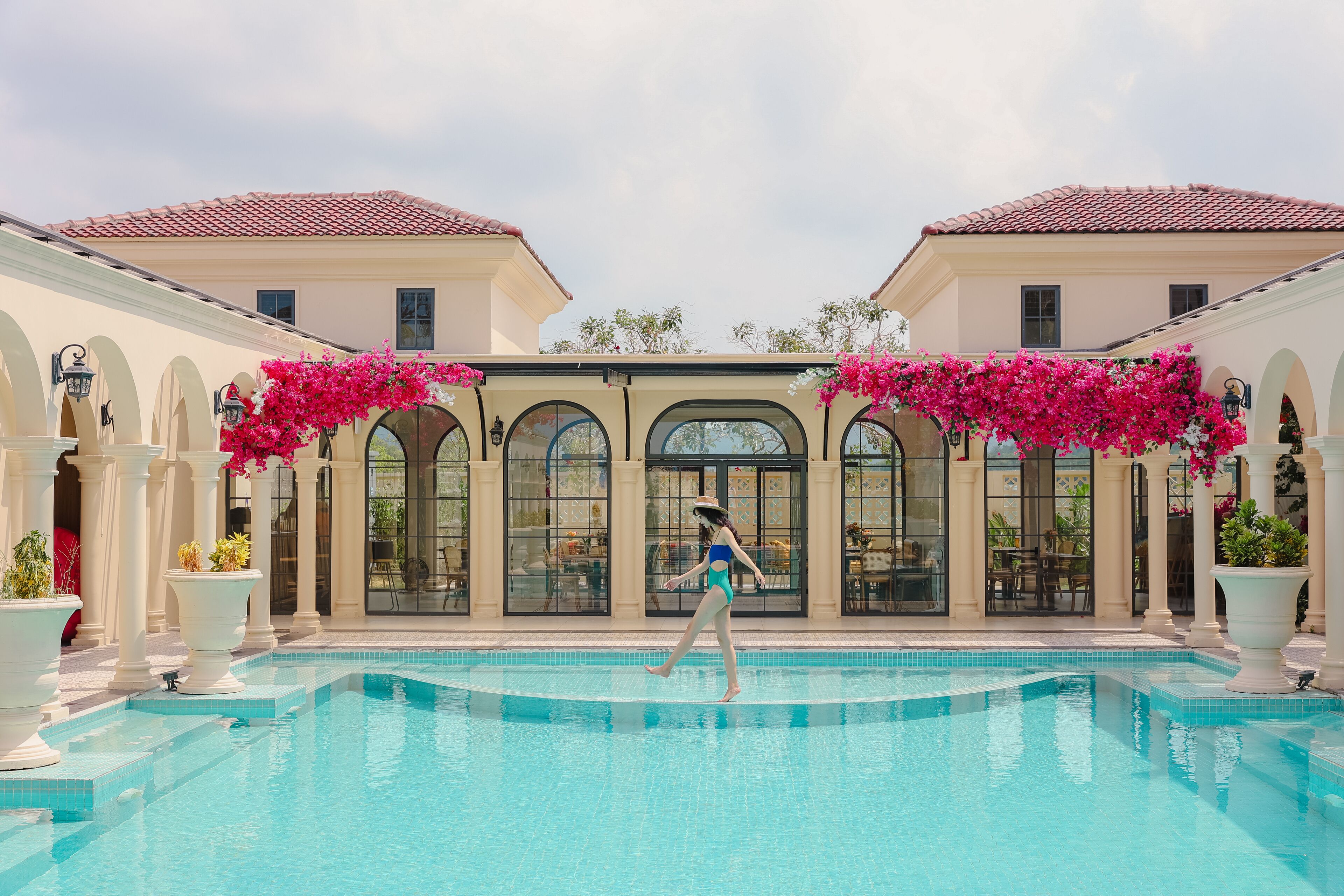 A swimming pool with a person walking, surrounded by columns and arches, and buildings with a tiled roof.