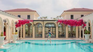 A swimming pool with a person walking, surrounded by columns and arches, and buildings with a tiled roof.