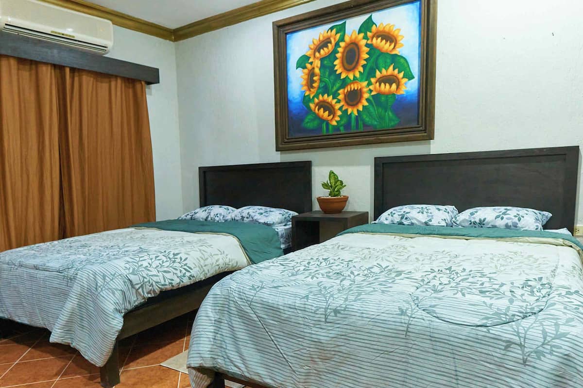 A hotel room with two beds, a potted plant, and a painting of sunflowers on the wall.