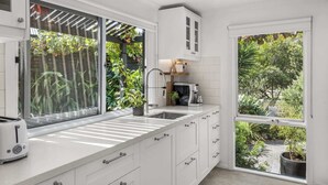 Eine moderne Küche mit weißen Schränken, einer integrierten Spüle und einer Kaffeemaschine. Große Fenster bieten Blick ins Grüne.