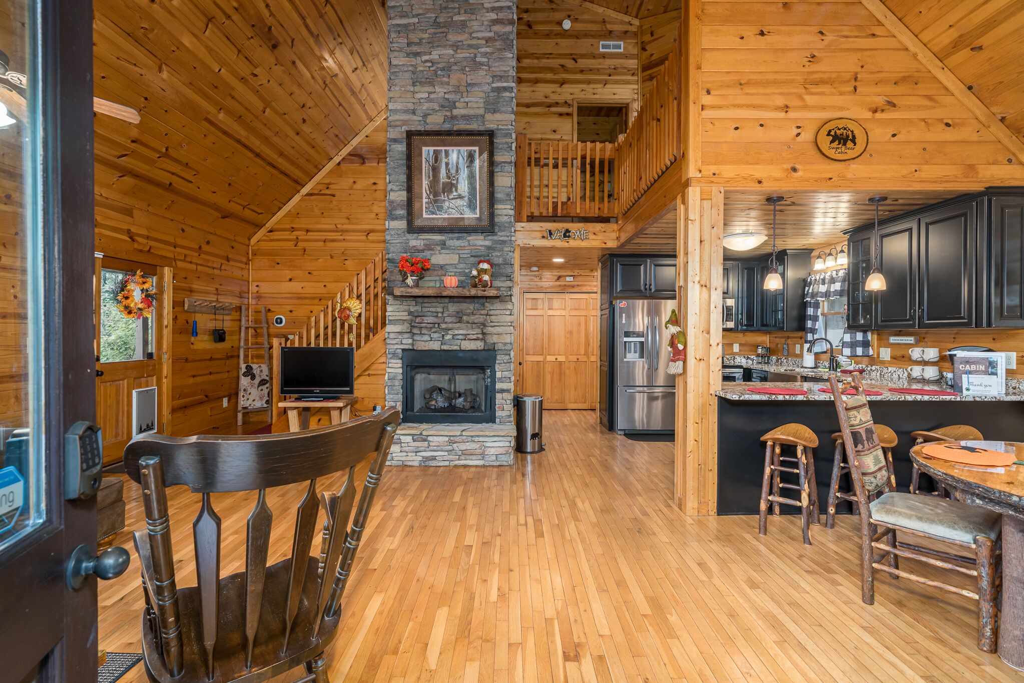 A log cabin interior with a stone fireplace, wooden dining area, and a kitchen with dark cabinets and stainless steel appliances.