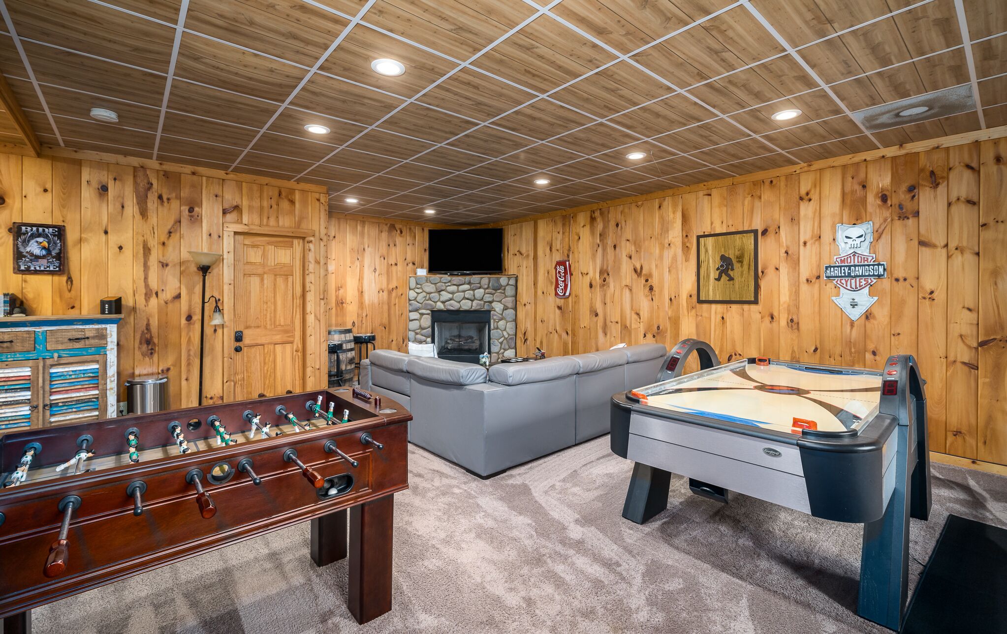 A cozy room with wooden walls, a foosball table, a pool table, a stone fireplace, and a sofa.