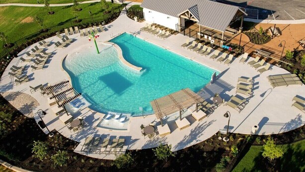 Ein Luftbild eines Resorts mit einem großen Schwimmbecken, umgeben von Liegestühlen und Sonnenbetten, einer überdachten Sitzgelegenheit und einem Gebäude mit geneigtem Dach.