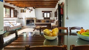 Uma sala de jantar rústica com uma mesa de madeira, cadeiras e um cesto de pão. Uma lareira é visível ao fundo, e uma geladeira está à direita.