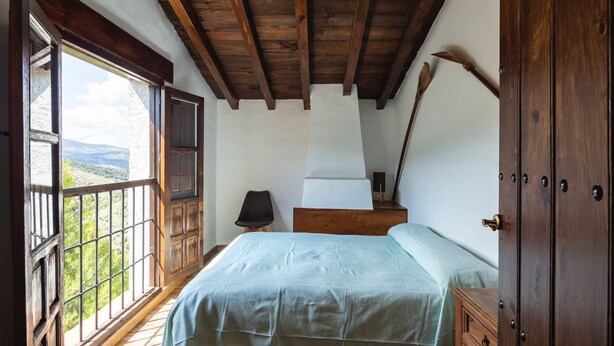 Quarto com teto de madeira, cama com colcha branca, mesa de cabeceira e janela com vista para o verde.