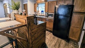 A kitchen with wooden cabinets, a black refrigerator, a sink, and a dining area with a glass table. - Rustic Bungalow - Rugged with a Touch of Class (Sulphur)