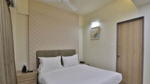 A neatly made bed with white linens, a wooden headboard, a bedside table with a lamp, and a door with a wooden panel. - hotel monville  (Vadodara)