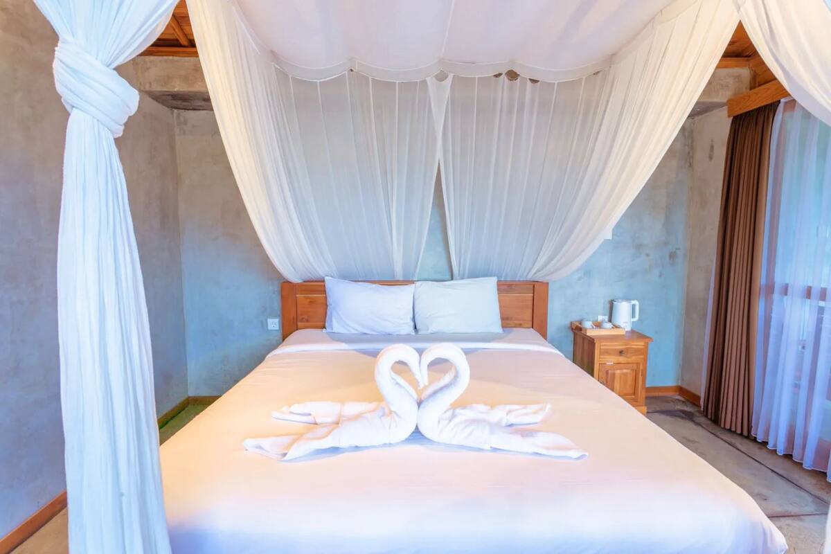 Une chambre d’hôtel avec un lit à baldaquin, une table de chevet en bois et une décoration en forme de cœur faite avec une serviette.
