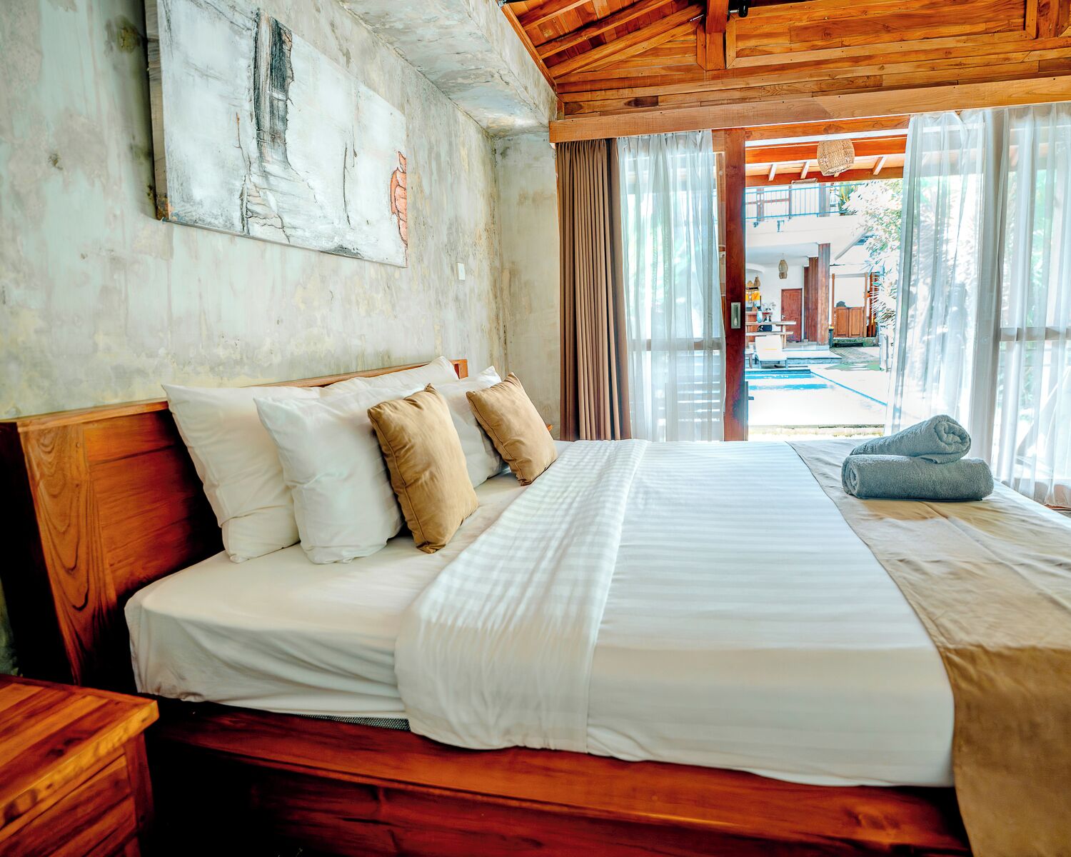 Una cama con ropa de cama blanca y beige, cabecera de madera y vista al área de una piscina exterior.