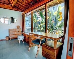 A room with a wooden desk, chairs, a rattan cabinet, and a large window with plants.