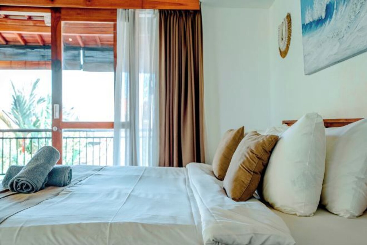 Ein Hotelzimmer mit Bett, Kissen, Blick durch ein Fenster und einem Gemälde an der Wand.