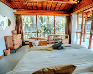 A room with a bed, wooden furniture, a window with a view of palm trees, and a balcony.