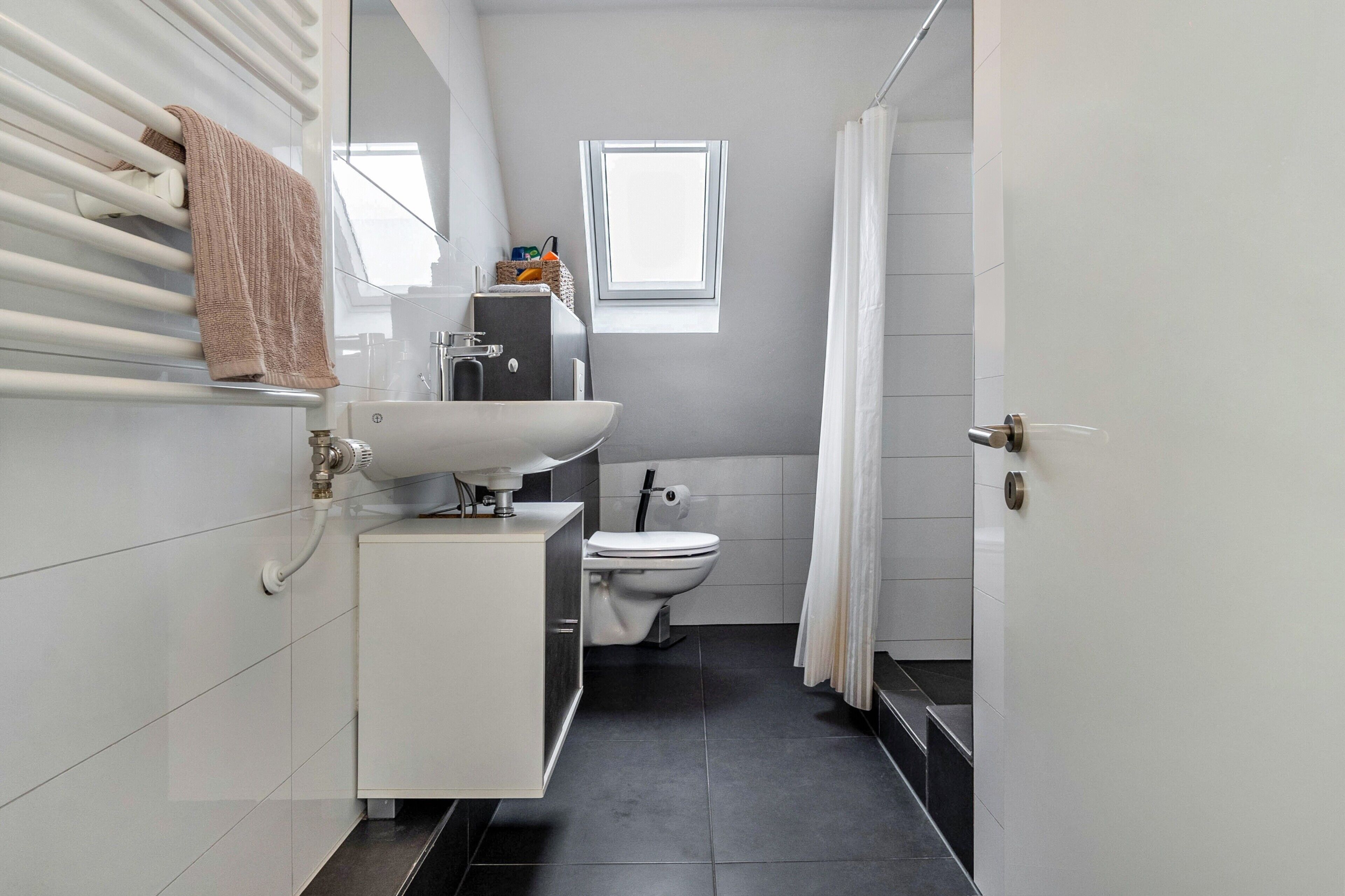 Un baño estrecho con lavamanos blanco, inodoro, toallero y ventana.